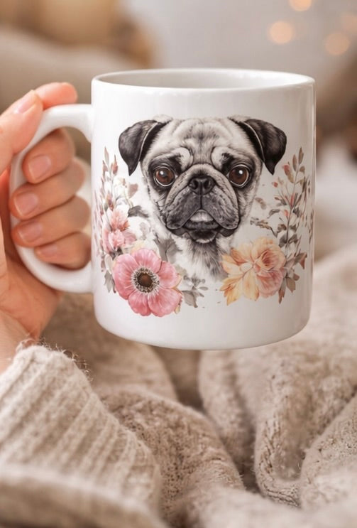 White mug with a pug and floral design held by a person wearing a knit sweater.